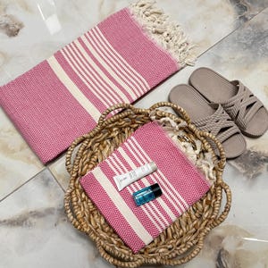 May include: Two pink and white striped towels, one in a wicker basket and one lying flat. The towel in the basket has a small blue bottle and a white tube of lotion on top of it. There are two brown sandals in the background.