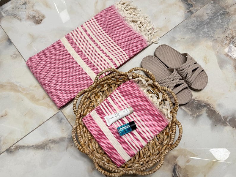 May include: Two pink and white striped towels, one in a wicker basket and one lying flat. The towel in the basket has a small blue bottle and a white tube of lotion on top of it. There are two brown sandals in the background.