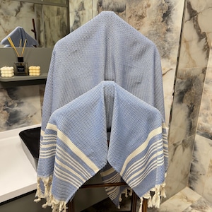 May include: Two blue and white striped Turkish towels with fringe edges. The towels are hanging on a wooden rack in a bathroom.