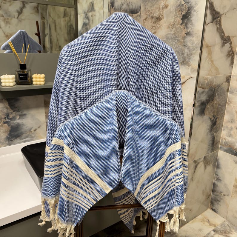 May include: Two blue and white striped Turkish towels with fringe edges. The towels are hanging on a wooden rack in a bathroom.