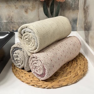 May include: Three rolled towels in beige, burgundy, and black patterns rest on a woven, circular placemat. The towels are stacked, showcasing their textures and colors. The background is a neutral-toned wall.