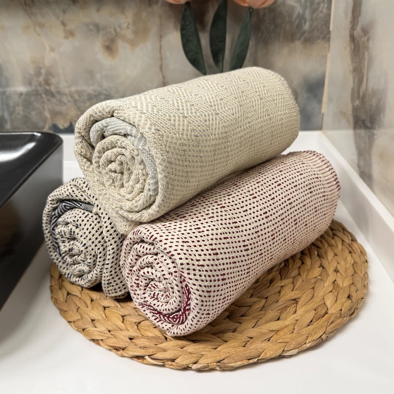 May include: Three rolled towels in beige, burgundy, and black patterns rest on a woven, circular placemat. The towels are stacked, showcasing their textures and colors. The background is a neutral-toned wall.