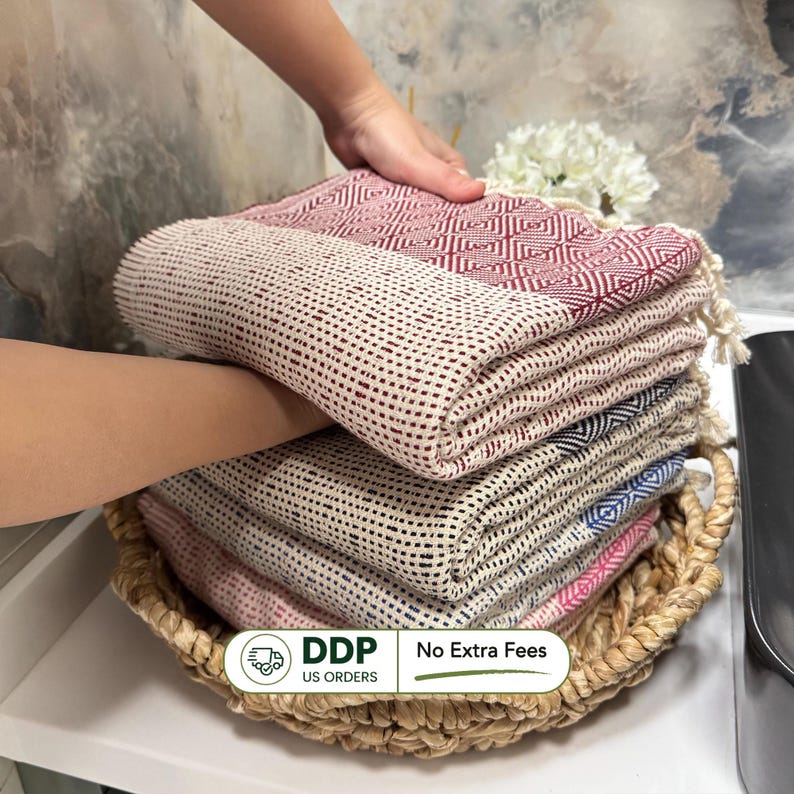 May include: A stack of colorful woven towels in a woven basket. The towels are in shades of red, pink, blue, and cream. The towels have a textured pattern and are neatly folded. The basket is made of natural fibers.