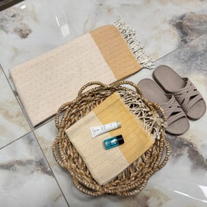 May include: A woven basket holds a yellow and cream-colored towel, a small tube of lotion, and a blue bottle. A larger cream and yellow towel with fringe and a pair of taupe sandals are nearby. The items are arranged on a tiled surface.