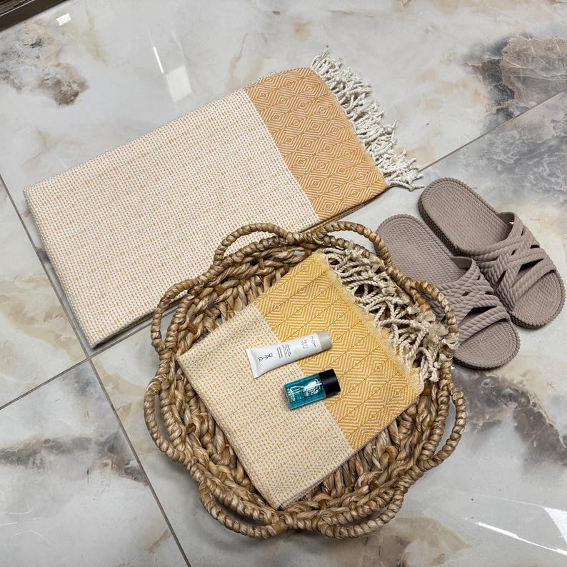 May include: A woven basket holds a yellow and cream-colored towel, a small tube of lotion, and a blue bottle. A larger cream and yellow towel with fringe and a pair of taupe sandals are nearby. The items are arranged on a tiled surface.
