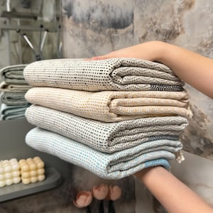 May include: A stack of folded, textured towels in various colors, including cream, beige, gray, and light blue. The towels have a woven pattern and are neatly arranged. The image is taken indoors, with a neutral background.