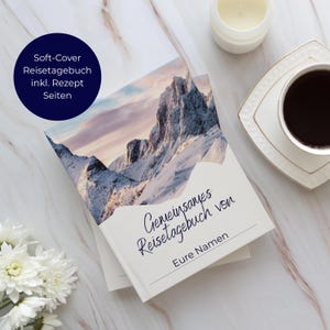 May include: A soft-cover travel journal featuring a mountain landscape on the cover. The journal is open, displaying the title "Gemeinsames Reisetagebuch von Eure Namen". A dark blue circle with white text reads "Soft-Cover Reisetagebuch inkl. Rezept Seiten". A cup of coffee and a candle are present.