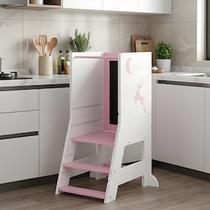 May include: A white wooden kitchen step stool with pink accents. The step stool has three steps and a safety bar. A pink deer and moon design is on the side. The step stool is designed to help children reach countertops.
