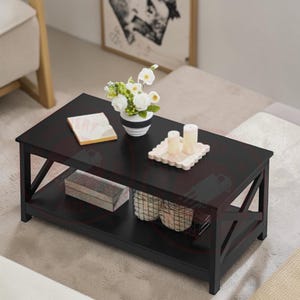 May include: A rectangular black coffee table with a lower shelf and X-shaped side supports. The top surface holds a white vase with white flowers, a small open book, and two cylindrical candles on a scalloped tray. A box and wire basket sit below.
