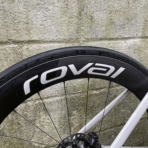 May include: A black bicycle wheel with a white "Roval" logo on the side. The wheel has a black rim and silver spokes. The wheel is mounted on a white bicycle frame.