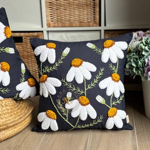 May include: Two navy blue throw pillows with embroidered white daisies and yellow centers. The daisies are arranged on green stems. The pillows are on a wooden surface, with a wicker basket and a shelf in the background.