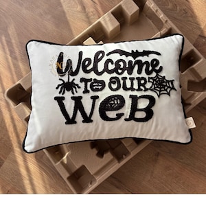 Op de afbeelding: Een rechthoekig wit kussen met zwart geborduurde tekst en Halloween-ontwerpen. De tekst luidt "Welcome to our web" met een spin, een pompoen en een spinnenweb. Het kussen heeft een zwarte rand.