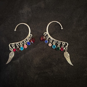May include: A pair of silver-toned ear cuffs. Each cuff features a curved design with a series of colorful, round gemstones in red, blue, pink, and green. Small, detailed wing charms dangle from the bottom of each cuff.