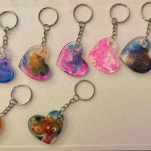 May include: A collection of ten heart-shaped resin keychains with various colorful abstract designs. The keychains are made with clear resin and feature swirls of blue, pink, purple, and orange colors. Some keychains have glitter embedded in the resin.