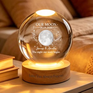Può includere: Una lampada a sfera in vetro trasparente su una base circolare in legno. La sfera presenta una grafica lunare incisa, stelle e il testo: OUR MOON 06.04.2026 James & Amelia The night our story began. La base in legno riporta: Love You Always Forever.