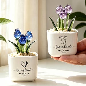 May include: Two white ceramic square planters containing glass flowers. One planter features blue glass tulips, a heart with a paw print, and the text "forever loved ZOE 2025". The other features purple glass tulips, a cat silhouette, and "forever loved LOLA 2025".