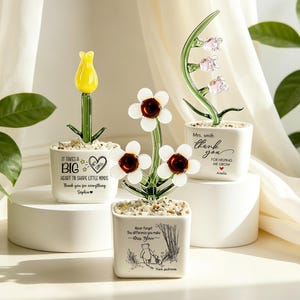 May include: Three square white ceramic pots containing glass flowers. One has a yellow tulip, one has three white flowers with brown centres, and one has pink lily-like flowers. Each pot features black printed text and decorative pebbles.