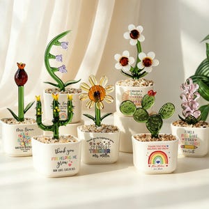 May include: A collection of six small white square ceramic pots, each containing a glass flower or cactus sculpture and decorative pebbles. The pots feature printed text and colourful graphics, including phrases like 'Thank you for helping me grow' and teacher names.