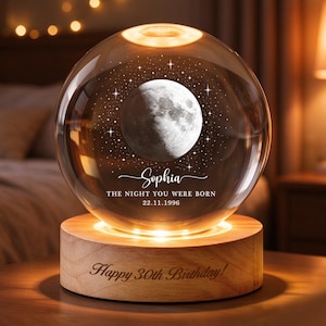 Puede incluir: Una lámpara de esfera de cristal transparente con un patrón grabado de luna y estrellas, el nombre Sophia y el texto THE NIGHT YOU WERE BORN 22.11.1996. La esfera descansa sobre una base de madera redonda grabada con Happy 30th Birthday.