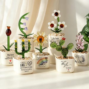 Op de afbeelding: Een verzameling van zes glazen bloem- en cactusbeeldjes in vierkante witte keramische potten gevuld met kleine steentjes. Elke pot is voorzien van gedrukte teksten en illustraties, waaronder diverse bedankjes en persoonlijke boodschappen.