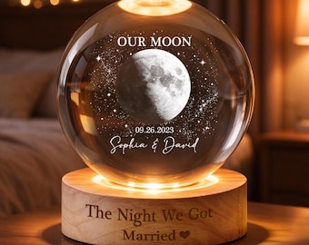 Op de afbeelding: Een bolvormige glazen lamp met een gegraveerd maan- en sterrenontwerp met de tekst "OUR MOON", "09.26.2023" en "Sophia & David". De voet is een cilindrische houten standaard gegraveerd met "The Night We Got Married" en een hartje.