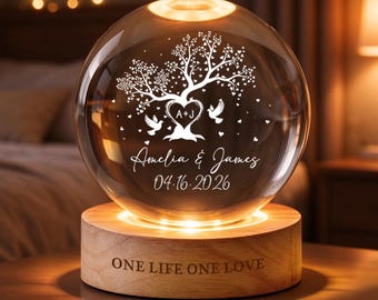 May include: A spherical glass lamp on a round wooden base. The glass features an etched tree with heart-shaped leaves, two birds, the initials A+J, the names Amelia & James, and the date 04.16.2026. The wooden base is engraved with ONE LIFE ONE LOVE.