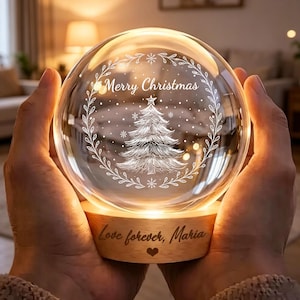 Könnte beinhalten: Ein kugelförmiges Glas-Nachtlicht mit einem eingravierten Weihnachtsbaum und dem Text Merry Christmas in einem Kranz. Das Licht steht auf einem runden Holzsockel, der mit dem Text Love forever, Maria und einem kleinen Herzsymbol graviert ist.