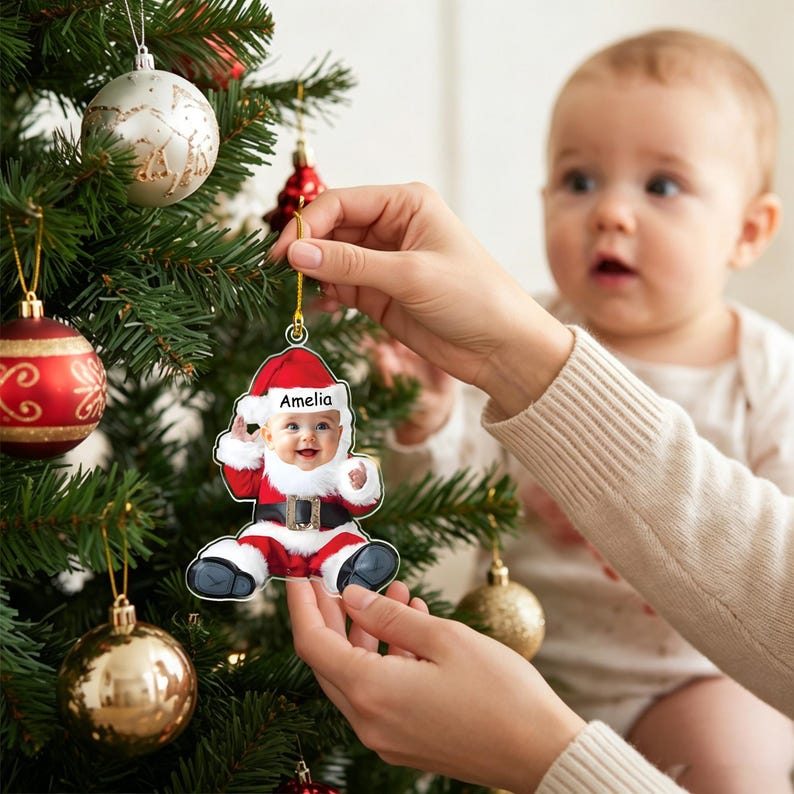 May include: A flat, custom-shaped Christmas tree ornament featuring a photograph of a baby dressed in a Santa Claus suit with a white fur-trimmed hat and coat. The name Amelia is printed in black text on the white brim of the Santa hat.