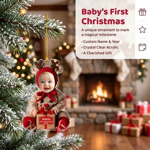 May include: A clear acrylic Christmas ornament featuring a photo of a baby wearing a brown reindeer hat and outfit, holding a small gift box labeled Amelia First Christmas 2026. The ornament hangs from a gold string on a pine tree branch.