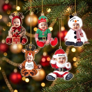 May include: Five personalized Christmas ornaments featuring photos of a baby in various costumes: a reindeer with a gift box labeled Amelia, an elf, a snowman, a reindeer with the text Amelia First Christmas 2026, and a Santa Claus with Amelia printed.