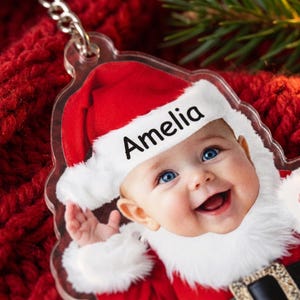 May include: A clear acrylic ornament featuring a printed image of a smiling baby wearing a red and white Santa hat with the name Amelia printed in black text on the white brim. The ornament includes a silver metal chain attachment at the top.