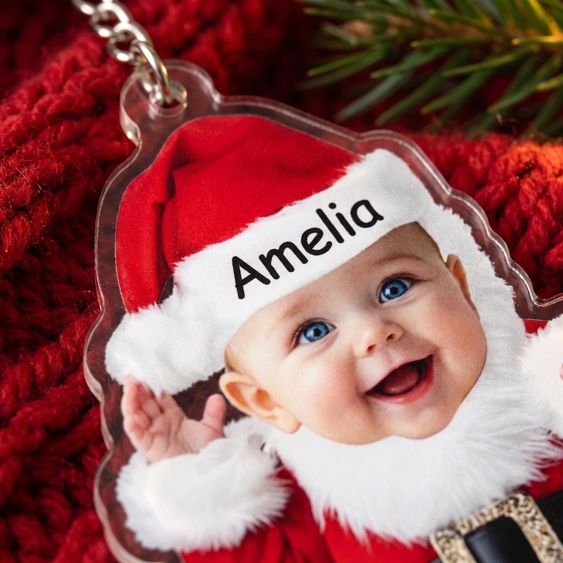May include: A clear acrylic ornament featuring a printed image of a smiling baby wearing a red and white Santa hat with the name Amelia printed in black text on the white brim. The ornament includes a silver metal chain attachment at the top.