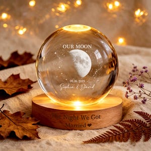May include: A clear glass sphere lamp on a round wooden base. The sphere features an etched moon, stars, the text "OUR MOON", "09.26.2023", and "Sophia & David". The wooden base is engraved with "The Night We Got Married" and a heart symbol.