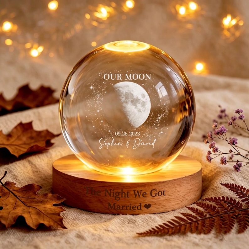 May include: A clear glass sphere lamp on a round wooden base. The sphere features an etched moon, stars, the text "OUR MOON", "09.26.2023", and "Sophia & David". The wooden base is engraved with "The Night We Got Married" and a heart symbol.