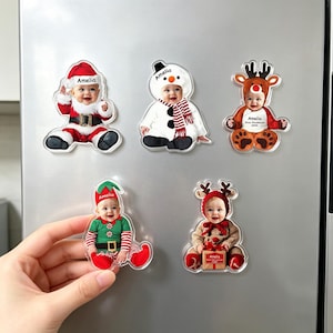 May include: Five custom acrylic magnets featuring a baby dressed in various holiday costumes including Santa Claus, a snowman, a reindeer, an elf, and a festive outfit with a gift. Each magnet displays the name Amelia printed on the clothing.