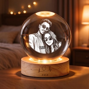 May include: A clear glass sphere lamp featuring an etched portrait of two people wearing sunglasses. The sphere rests on a round wooden base engraved with the letters j and b separated by a heart symbol and the date 5.12.18 below.