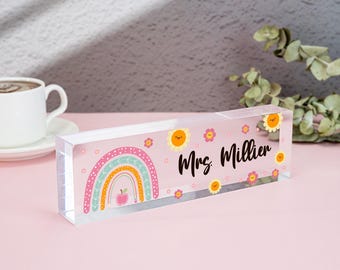 May include: A clear acrylic desk nameplate with a pink background and a rainbow, flowers, and a red apple design. The nameplate reads "Mrs. Miller".