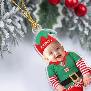 May include: A clear acrylic Christmas ornament shaped like an elf, featuring a photograph of a baby wearing a green and red elf costume with striped sleeves. The name Amelia is printed in white on the red elf hat. It hangs from a gold cord.