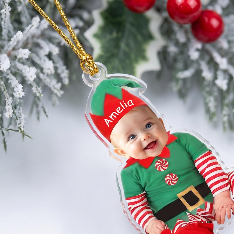 May include: A clear acrylic Christmas ornament shaped like an elf, featuring a photograph of a baby wearing a green and red elf costume with striped sleeves. The name Amelia is printed in white on the red elf hat. It hangs from a gold cord.