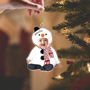 May include: A flat, custom-shaped Christmas ornament featuring a photograph of a baby dressed in a white snowman costume with a black hat, orange nose, and a red and white striped scarf. The name Amelia is printed in black text on the snowman hood.