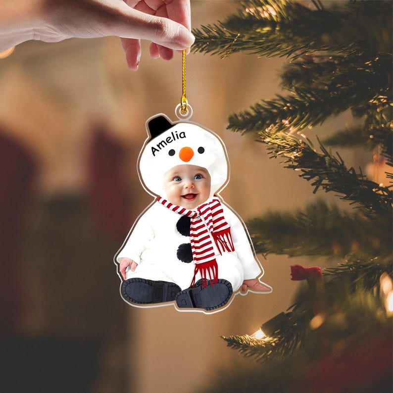 May include: A flat, custom-shaped Christmas ornament featuring a photograph of a baby dressed in a white snowman costume with a black hat, orange nose, and a red and white striped scarf. The name Amelia is printed in black text on the snowman hood.
