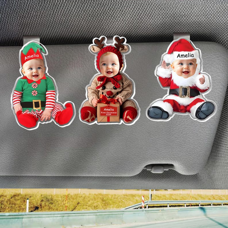 May include: Three custom car visor ornaments featuring a baby with the name Amelia. The left ornament shows the baby as an elf, the center as a reindeer holding a gift box labeled Amelia First Christmas 2026, and the right as Santa Claus.