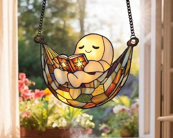 May include: Stained glass lamp featuring a smiling, glowing figure reading a book while relaxing in a hammock. The hammock is made of colorful glass pieces in shades of orange, yellow, and brown. The lamp hangs from a chain.