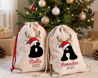 Puede incluir: Dos bolsas de regalo beige con cordones rojos. Cada bolsa presenta una letra negra, un gorro de Papá Noel con cuernos de reno y los nombres "Bella" y "Amadar" con el año "2025". Un árbol de Navidad está en el fondo.