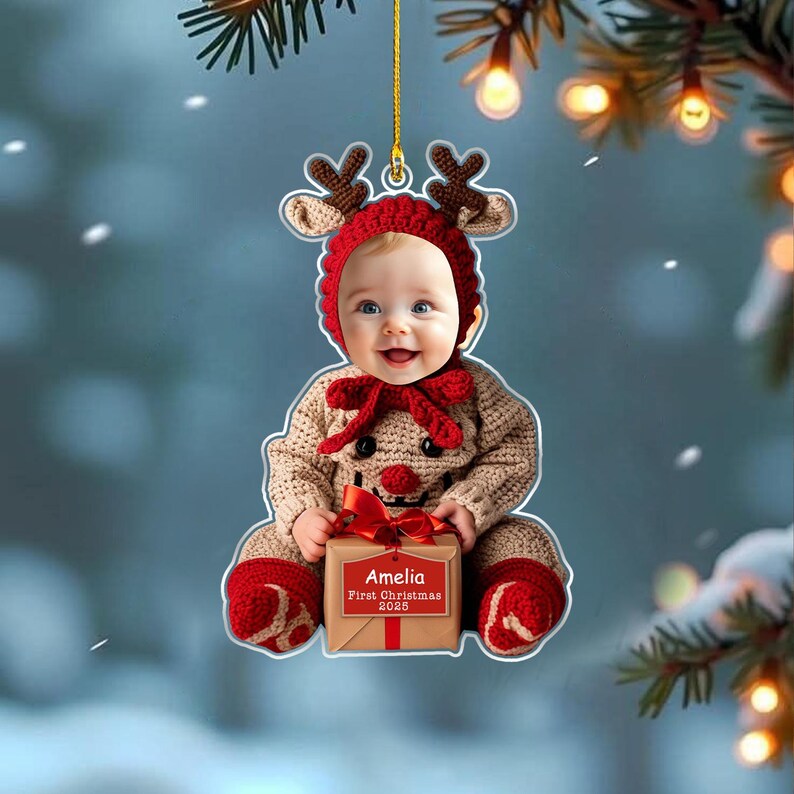 May include: A flat acrylic Christmas ornament featuring a photo of a baby wearing a crocheted reindeer hat and outfit. The baby holds a gift box with a red ribbon and a tag that reads Amelia First Christmas 2025. The ornament hangs by a gold cord.