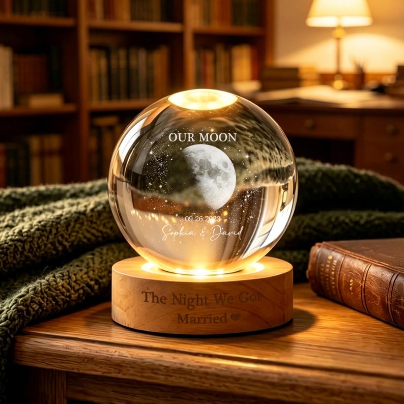 May include: A clear glass sphere lamp featuring an internal laser-etched moon and star design, with the text "OUR MOON", "09.26.2023", and "Sophia & David". It rests on a round wooden base engraved with "The Night We Got Married".