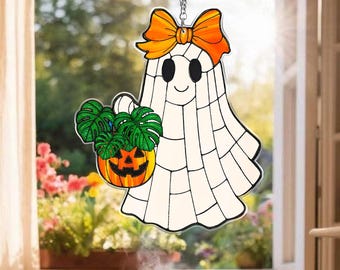 May include: A Halloween-themed window hanging featuring a smiling ghost with an orange bow and a jack-o'-lantern holding green plants. The ghost is white with black eyes and a black outline. The window hanging is suspended by a silver chain.