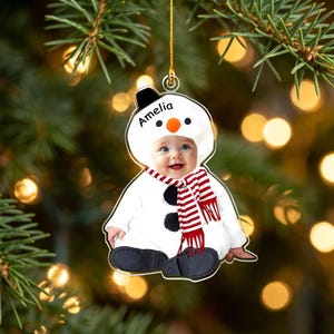 May include: A custom acrylic Christmas ornament featuring a photograph of a baby dressed in a white snowman costume with a black hat, orange nose, and red and white striped scarf. The name Amelia is printed in black text on the snowman hood.