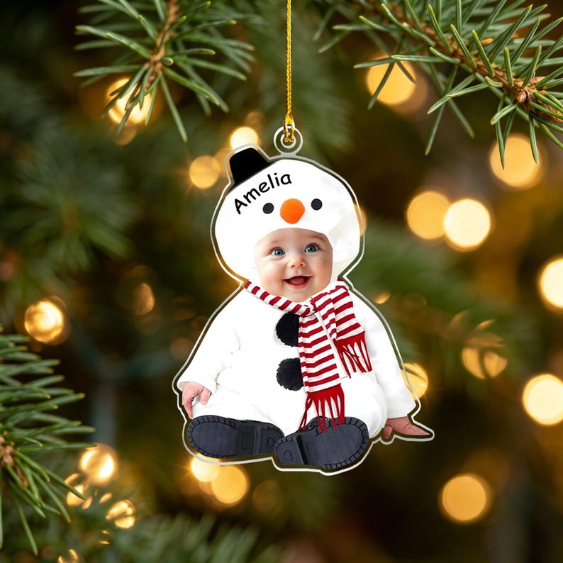 May include: A custom acrylic Christmas ornament featuring a photograph of a baby dressed in a white snowman costume with a black hat, orange nose, and red and white striped scarf. The name Amelia is printed in black text on the snowman hood.