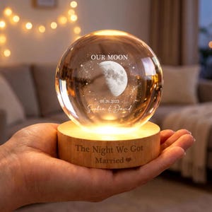 May include: A clear glass sphere night light featuring an internal laser-etched moon, stars, the text "OUR MOON", "09.26.2023", and "Sophia & David". The sphere rests on a round wooden base engraved with "The Night We Got Married".